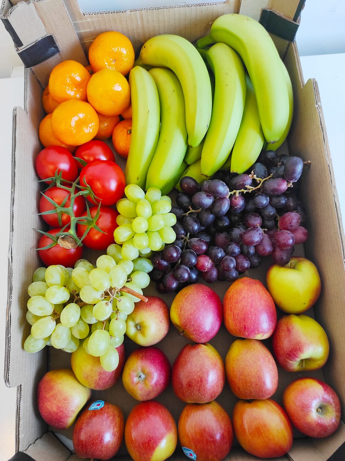 Box filled with assorted fruits including bananas, apples, grapes, tomatoes, and mandarins.